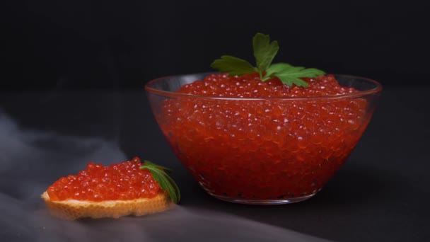 Spectacular presentation of traditional holiday Russian delicacy - red salmon caviar in glass bowl and on piece of bread decorated with fresh parsley leaf, clouds of smoke around, black background — Stock Video