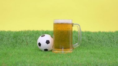 Bir kupa soğuk, ferahlatıcı köpük birası yeşil çimlerin üzerinde duruyor. Yapay çimenler, sarı arka plan. Küçük oyuncak futbol topu yuvarlanıyor. İnsan susuzluğunu gidermek için bardağı alır ve yerine koyar.