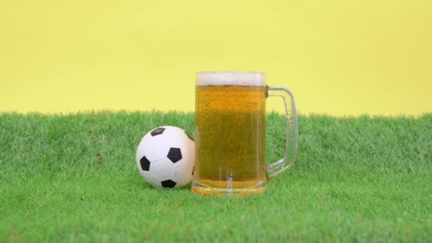 Taza de cerveza de espuma refrescante frío se encuentra en la hierba verde de césped artificial, fondo amarillo. Pequeña pelota de fútbol de juguete se despliega. El hombre toma la copa para saciar su sed y la devuelve — Vídeo de stock
