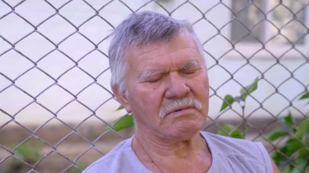 Un bătrân cu mustață gri și păr dozat, sprijinindu-se pe un gard de plasă metalică. În aer liber, zi de vară — Videoclip de stoc