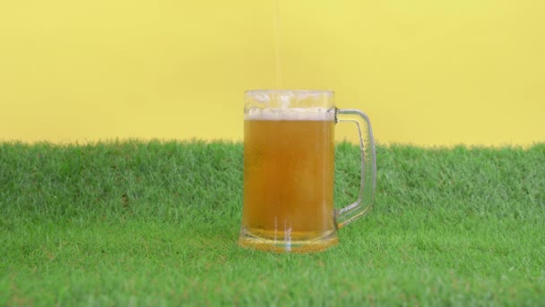 Studené osvěžující pěnové pivo se nalévá do velkého hrnku, stojany na zelené trávě z umělého trávníku, žluté pozadí, výhled zepředu. Malé hračky fotbal míč roll out. — Stock video