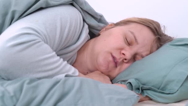 Jonge blonde vermoeide vrouw in pyjama slaapt, mompelt en snurkt in bed onder warme deken thuis. Gezondheid en ademhalingsproblemen, somnambulisme — Stockvideo