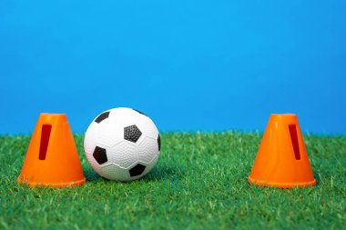 İki plastik koni bir futbol kalesini temsil ediyor, suni çimlerin yeşil çimlerinde küçük bir futbol topu, mavi arka plan, ön görüş, yakın çekim.