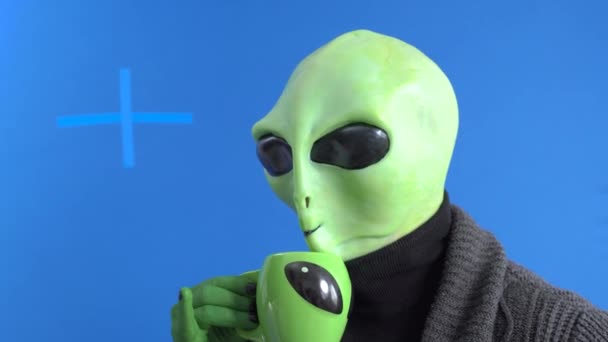 Man in alien costume and warm knitted jacket mannerly drinks hot drink from themed mug with his little finger sticking out against blue chromakey background — Stock Video