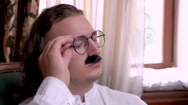 Un joven extraordinario con el pelo largo y el bigote grueso en camisa blanca baja levemente las gafas en su nariz y valora algo con mirada altiva hacia abajo — Vídeo de stock