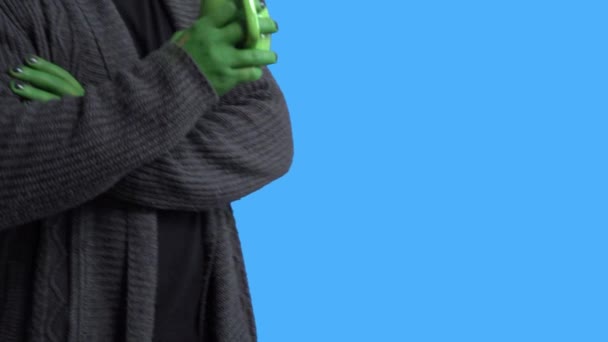 Professional actor in alien costume and cozy home outfit comes into the frame with funny mug and sits down to drink hot beverage against blue chromakey background — Stock Video