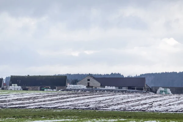 耕作可能な土地の雪のビット 酪農場納屋 白と青の乾草およびサイレージの納屋の近くのロールします ヨーロッパの冬の始まり — ストック写真