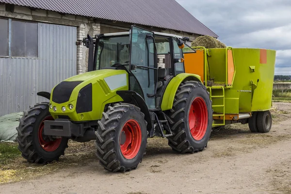 Tractor with a trailer for mixing and distribution of feed for cows. Behind the tractor is a barn and fields. Necessary equipment for a dairy farm.