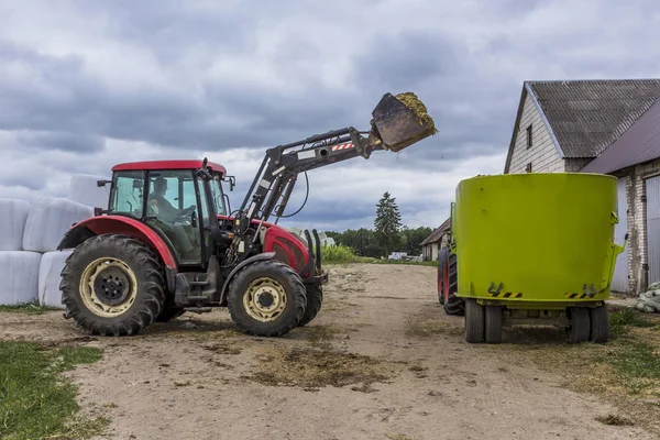 A tractor with a front end loader loads feed into an animal feed distributor for cows. Background barns and bales of silage. Dairy farm equipment.