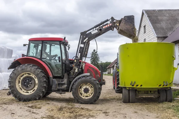 A tractor with a front end loader loads feed into an animal feed distributor for cows. Background barns and bales of silage. Dairy farm equipment.