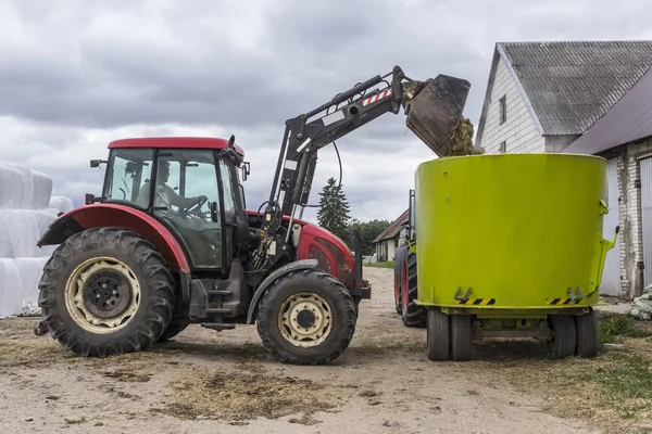 A tractor with a front end loader loads feed into an animal feed distributor for cows. Background barns and bales of silage. Dairy farm equipment.