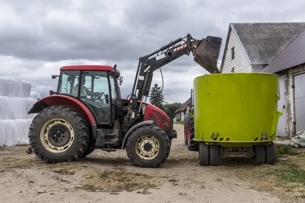 A tractor with a front end loader loads feed into an animal feed distributor for cows. Background barns and bales of silage. Dairy farm equipment.