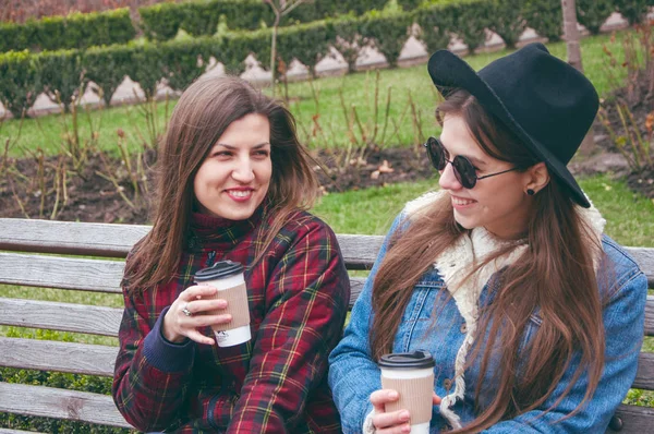 Beautiful European girls drink coffee and chat on the bench against the backdrop of the city — Stock Photo, Image