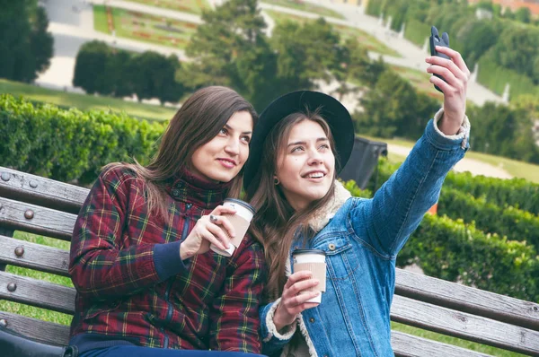 Hermosas chicas europeas toman café y hacen selfie en el banco contra el telón de fondo de la ciudad — Foto de Stock