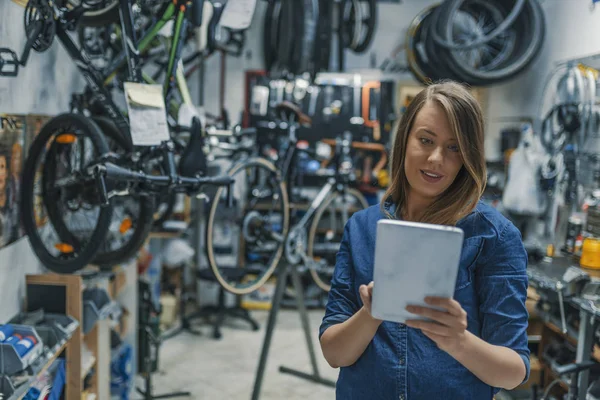 Female mechanic at work. Using a tablet while fixing a bicycle. Female bicycle repair technician using digital tablet in bicycle shop. Verify the checklist. Mechanic woman checking something on a tablet-pc and checklist.