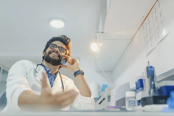 Smiling doctor talking on mobile phone at hospital. Handsome young doctor in white coat and eyeglasses is talking on the mobile phone while working in office