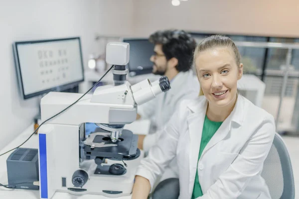 研究室で活躍する白い制服の若手 研究室で同僚の背景にカメラを見て魅力的な笑みを浮かべて 男性科学 — ストック写真