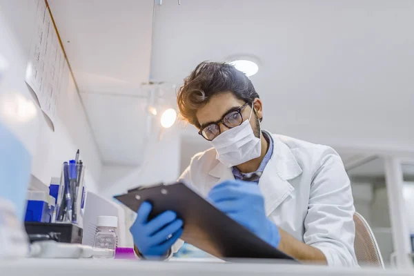 年轻的男性实验室技术员在一些样品上工作 并在剪贴板上做笔记 研究员在化学实验室的办公桌上写笔记 专业实验室技术员做笔记 — 图库照片