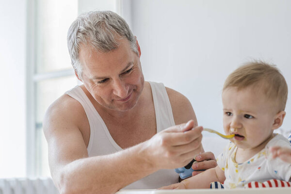 Отец и сын. Отец кормит маленького сына и веселится. Отец кормит шестимесячного ребенка ложкой. Мужчина проводит время со своим сыном. Отец мальчика занимается воспитанием ребенка. Отец кормит своего сына
. 