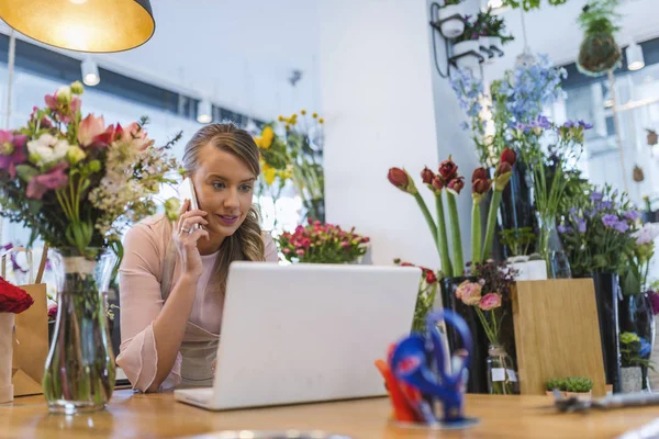 Happy florist using technologies at flower shop. Florist taking orders from the internet. Florist using interface of online flower shop. Florist talking on mobile phone while using laptop at her flower shop