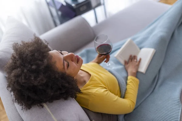 Relaxed young woman sitting on sofa with book and glass of red wine