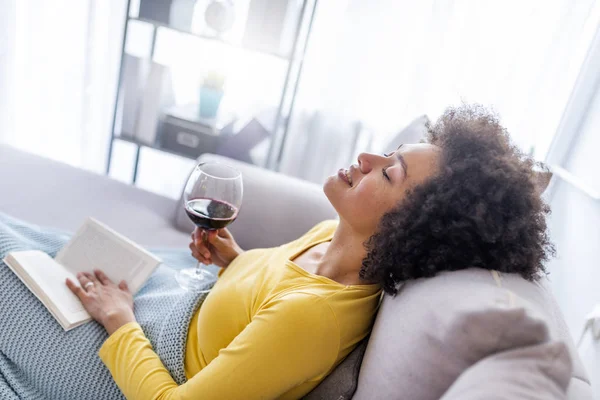 Relaxed young woman sitting on sofa with book and glass of red wine