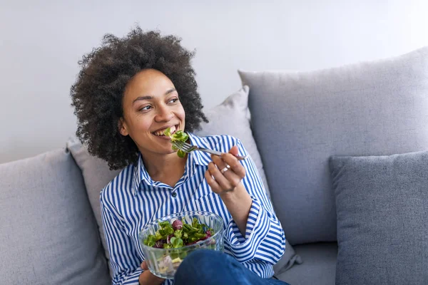 Beautiful African American woman eating salad on sofa in living room