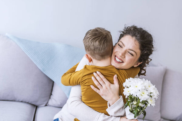 Mother and little son with bouquet of flowers hugging on sofa