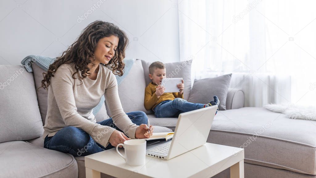 Mother working on laptop at home while son using tablet on sofa