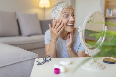 Aynada kendine bakan kadın. Güzel yaşlı kadın ayna tutan ve evde yüz kremi uygulayarak. Yaşlı kadın anti-kırışıklık kremi uygulayarak. 
