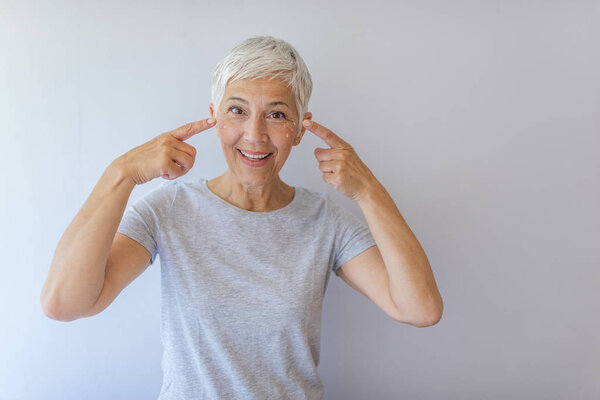 Mature woman applying anti-wrinkle face cream over gray background.