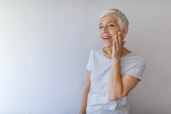 Mature woman applying anti-wrinkle face cream over gray background.