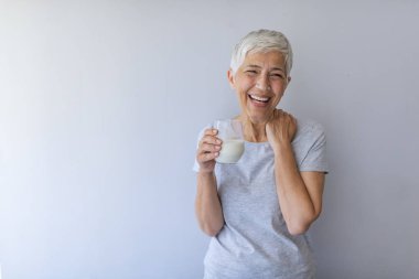 Evde bir bardak süt tutan kıdemli kadın. Orta yaş kadın ayakta mutlu bir yüz ile taze süt bir bardak içme ve dişleri gösteren kendinden emin bir gülümseme ile gülümseyerek