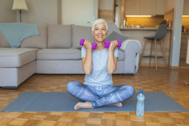 Halter çalan kıdemli kadının portresi. Üst düzey kadın tv önünde fitness exercices yapıyor. Happy yaşlı kadın dumbbells ile fitness eğitimi yapma