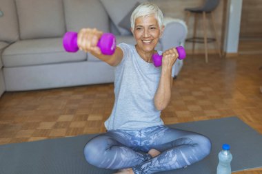 Aktif kıdemli emekli kadın evde ağırlıkları ile egzersiz. Dumbbell tutan gülümseyen kıdemli kadın portresi. İyi bir sağlığa uzanıyor. Evde Olgun Kadın Egzersiz. 