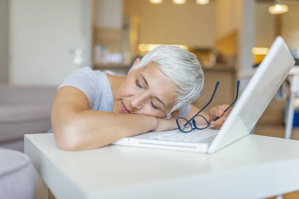 Portrait of a tired mature businesswoman sitting at the table with laptop computer while holding cup of coffee and sleeping at home. Senior woman Sleeping on Desk