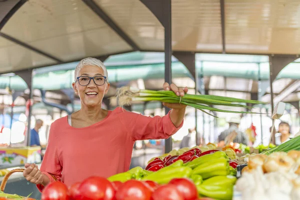Mature Female Customer Shopping At Farmers Market Stall. Woman shopping at local Farmers market. Mature woman buying vegetables.