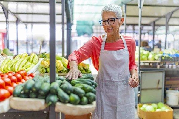 Female Stall Holder At Farmers Fresh Food Market. Friendly senior woman tending an organic vegetable stall at a farmer\'s market and selling fresh vegetables. Greengrocer selling organic fresh agricultural product