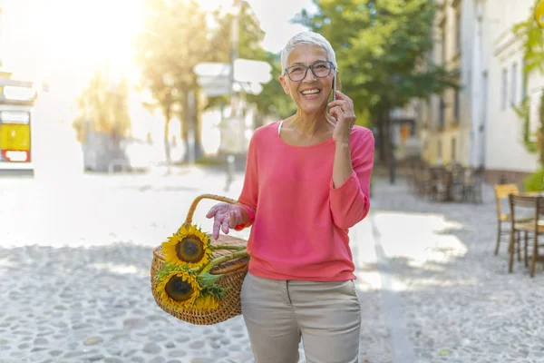 Şehirde açık havada yürürken akıllı telefon kullanan zarif yaşlı kadın. Emeklilikte iş hayatı kavramı. Mutlu yaşlı kadın sokakta yürüyüş ve akıllı telefon çağırıyor