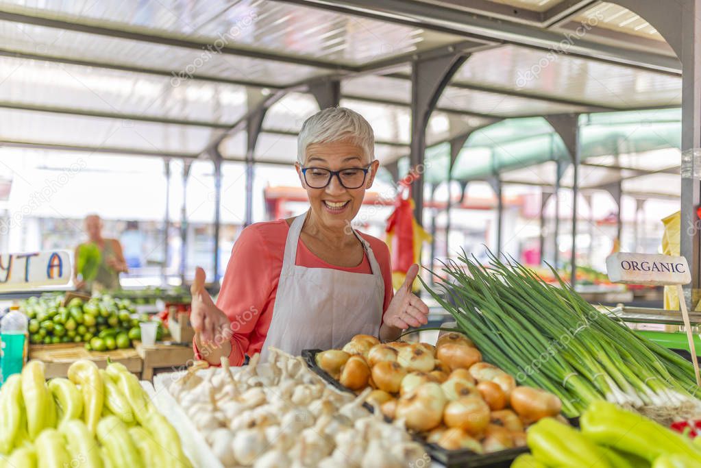 Greengrocer selling organic fresh agricultural product at farmer market. Friendly senior woman tending an organic vegetable stall at a farmers market. 