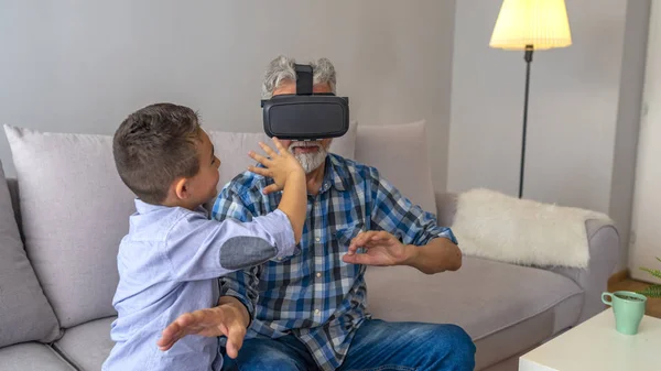 Grandson try to demonstrate how to use VR machine with grandfather in school holidays period. Family relationship concept. Portrait of excited senior man using VR glasses sitting on sofa at home with laughing grandson beside him