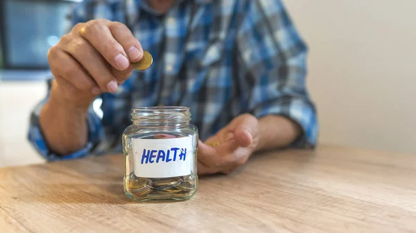 Saving money for health. Man Dropping Coins Into Glass Jar Labelled Health. Senior man putting Coins in glass jar with Health label. Responsible investing