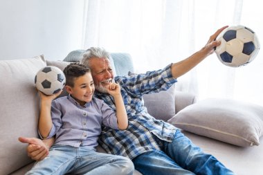 Dedesi ile kanepede küçük çocuk, bir futbol oyunu için tezahürat ve bir futbol topu tutan. Dedesi ile kanepede küçük çocuk, bir futbol oyunu için tezahürat ve evde bir futbol topu tutarak... 