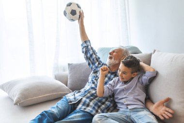 Heyecanlı torun ve futbol maçı izlerken kıdemli adam. Overjoyed dedesi ve çocuk birlikte futbol maçı izlerken, bir futbol oyunu için tezahürat ve bir futbol topu tutarak 