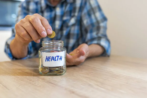 Saving money for health. Man Dropping Coins Into Glass Jar Labelled Health. Senior man putting Coins in glass jar with Health label. Responsible investing