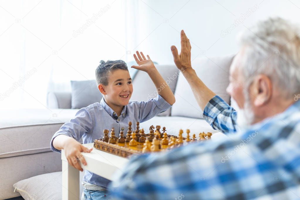 Happy grandfather plays chess with small grandson. Development of logic and strategic thinking.  Young boy is playing chess with his grandfather at home. Happy boy grandson playing chess with old senior man 