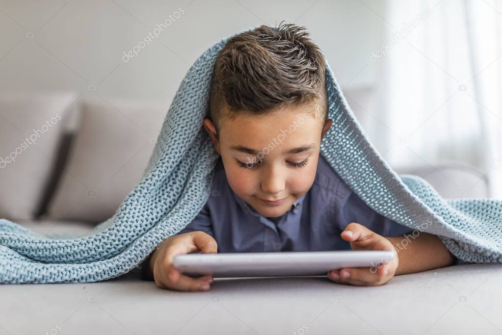 Little boy with digital tablet sitting on sofa, on home interior background. Boy playing with digital tablet. Little  boy sitting on sofa and playing game on digital tablet. 