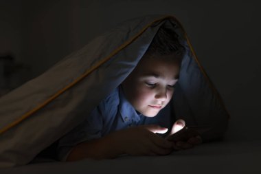 Battaniyenin altında cep telefonu olan küçük çocuk. Akıllı telefonla yatakta yatan çocuk. Genç yatağında akıllı telefondan e-posta göndererek. 7 yaşındaki çocuk çocuk yatakta gece akıllı telefon kullanarak