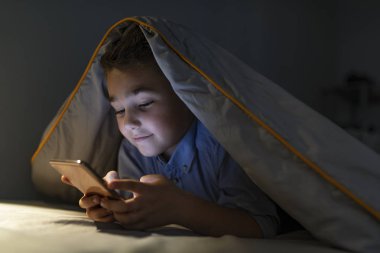 Küçük çocuk geceleri çizgi film izliyor. Küçük çocuk, karanlıkta yatarken, akıllı telefonla oynuyor. Battaniyenin altında yatan ve cep telefonuyla oynayan çocuk portresi