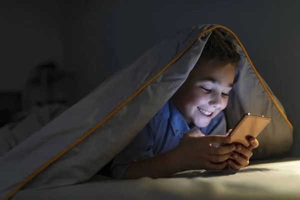 Kid boy playing smartphone lying on a bed at night. 7 years old child boy using smartphone at night in bed. Boy in bed texting on smartphone. Child with smart phone